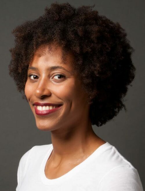 A Black woman wearing a white crew neck shirt, red lipstick, and an Afro hairstyle, smiles at the camera.
