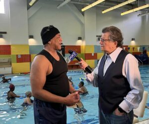 Reporter interviews man is swimsuit standing in front of an indoor swimming pool