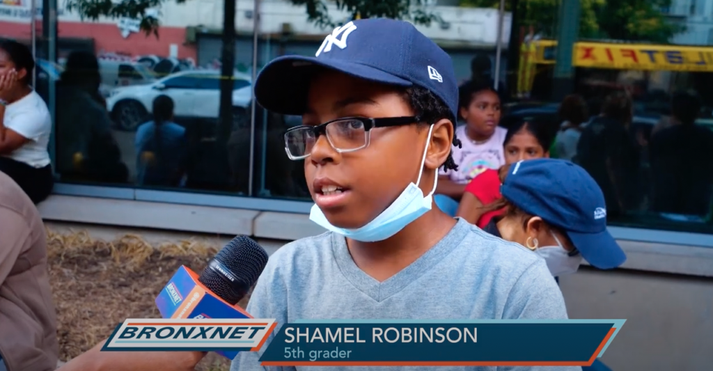 An elementary aged Black boy, wearing a baseball cap, pulled down face mask, and a light blue shirt, gives a news interview in front of a school building.