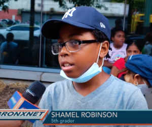 An elementary aged Black boy, wearing a baseball cap, pulled down face mask, and a light blue shirt, gives a news interview in front of a school building.