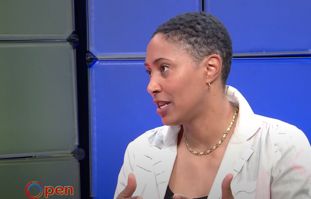 Black woman with short hair wearing a gold chain and light colored blazer being interviewed.