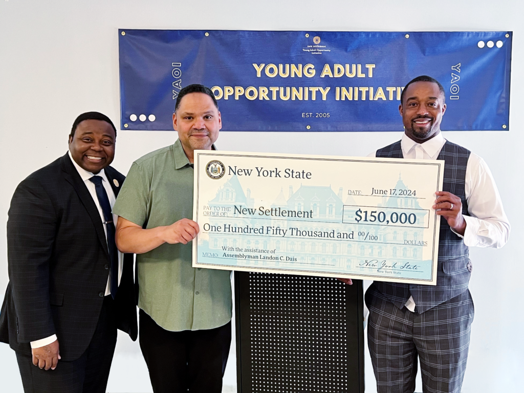 The image shows three men standing in front of a banner that reads "Young Adult Opportunity Initiative." They are holding an oversized check from New York State for $150,000 made out to "New Settlement." The date on the check is June 17, 2024. The memo line notes the assistance of Assemblyman Landon C. Dais. The man in the middle is holding the check, while the two men on either side are smiling at the camera.